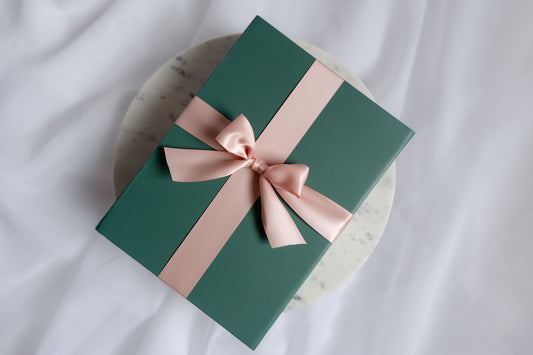 A dark green gift box wrapped with a wide pale pink satin ribbon and bow, placed on a round white marble coaster over a white fabric background.