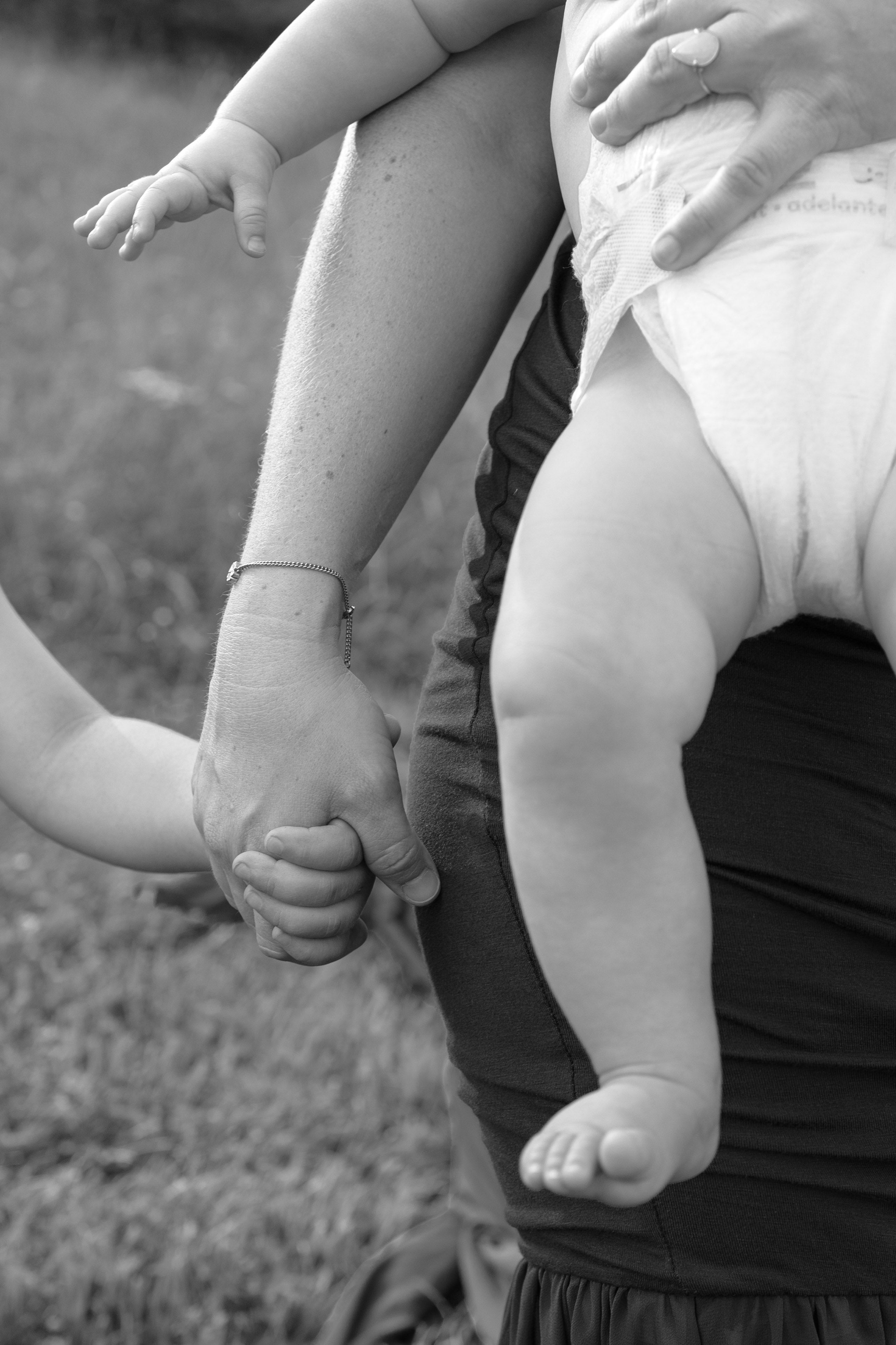 Black and white close-up of a baby in a white diaper held by a mama in the outdoors, with the baby's foot resting by her hand and her toddlers.