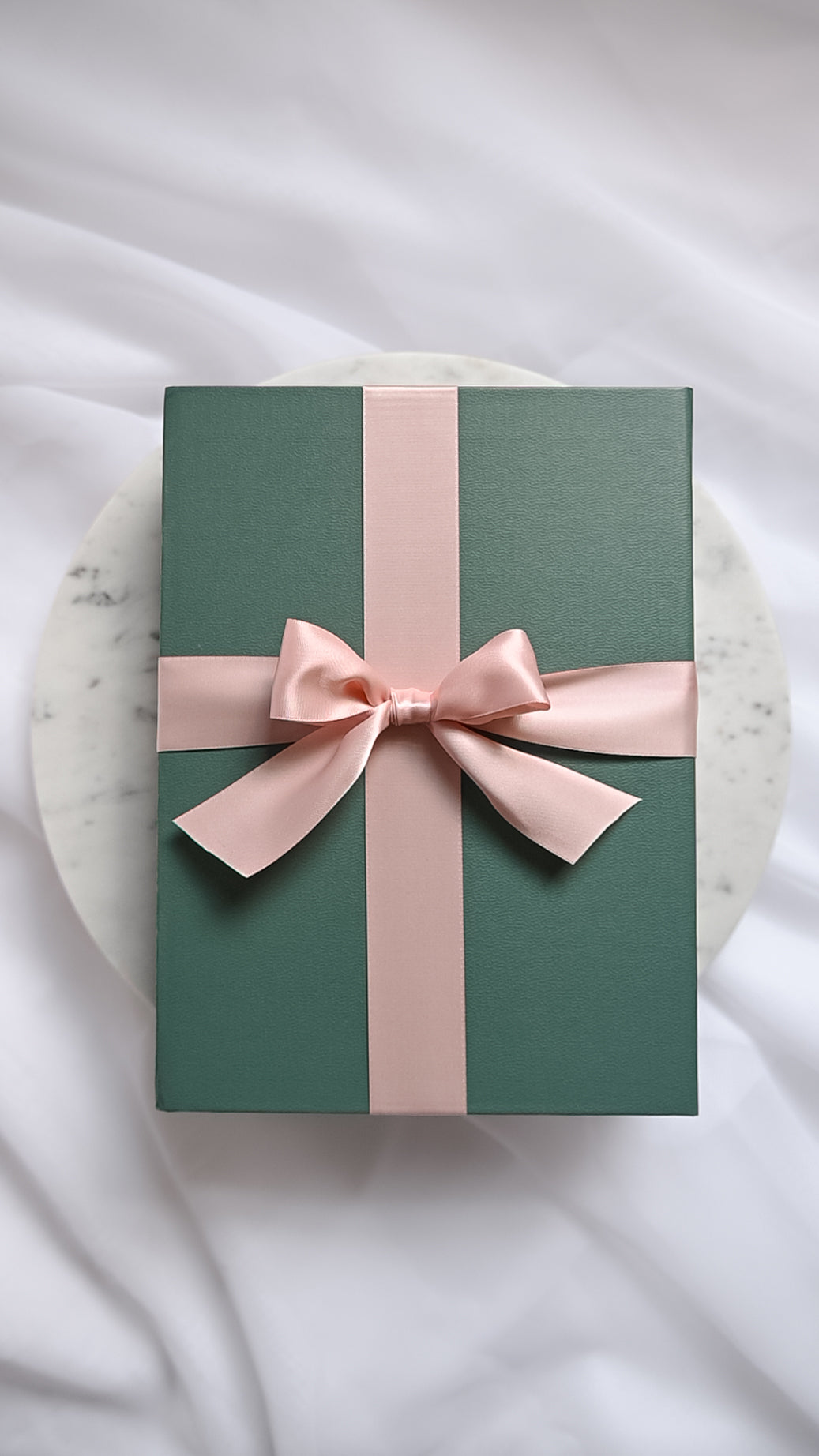 A dark green gift box wrapped with a light pink satin ribbon and bow, placed on a round white marble plate over a white fabric background.