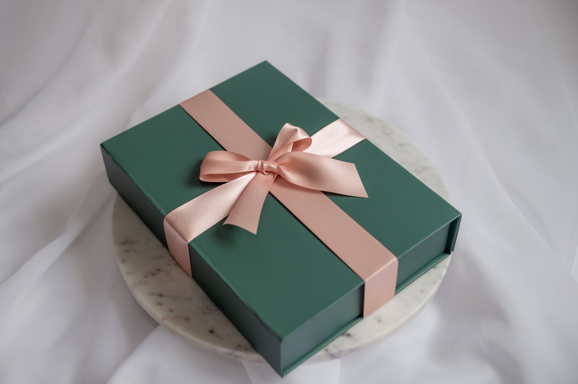 A dark green rectangular gift box tied with a wide blush pink satin ribbon and bow, displayed on a round white marble plate against a soft white fabric backdrop.