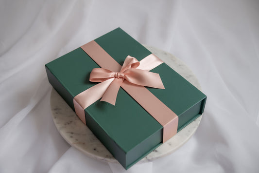 A dark green rectangular gift box tied with a wide blush pink satin ribbon and bow, displayed on a round white marble plate against a soft white fabric backdrop.