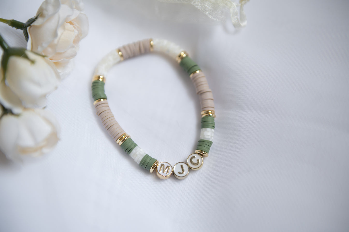 A clay-beaded personalized bracelet with sage green, cream, and beige beads, accented with gold spacer rings and letter beads spelling baby's initials, displayed on a white fabric background with soft white flowers in the corner.