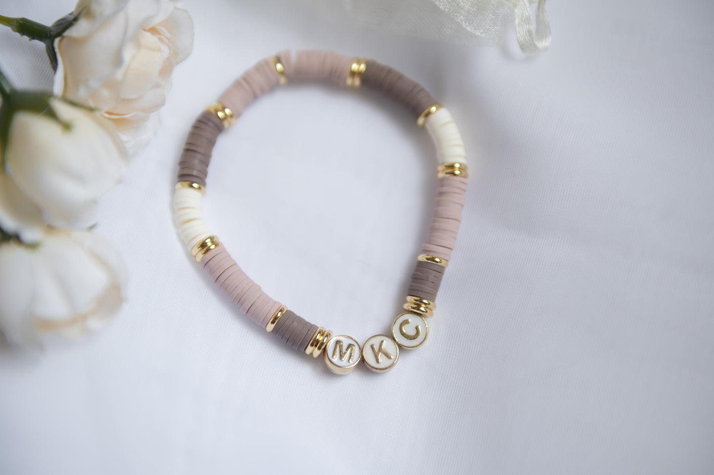 A clay-beaded bracelet with alternating dusty pink, taupe, and cream beads, accented with gold spacer rings and three round gold letter beads spelling baby's initials, displayed on a white surface with soft white flowers in the corner.