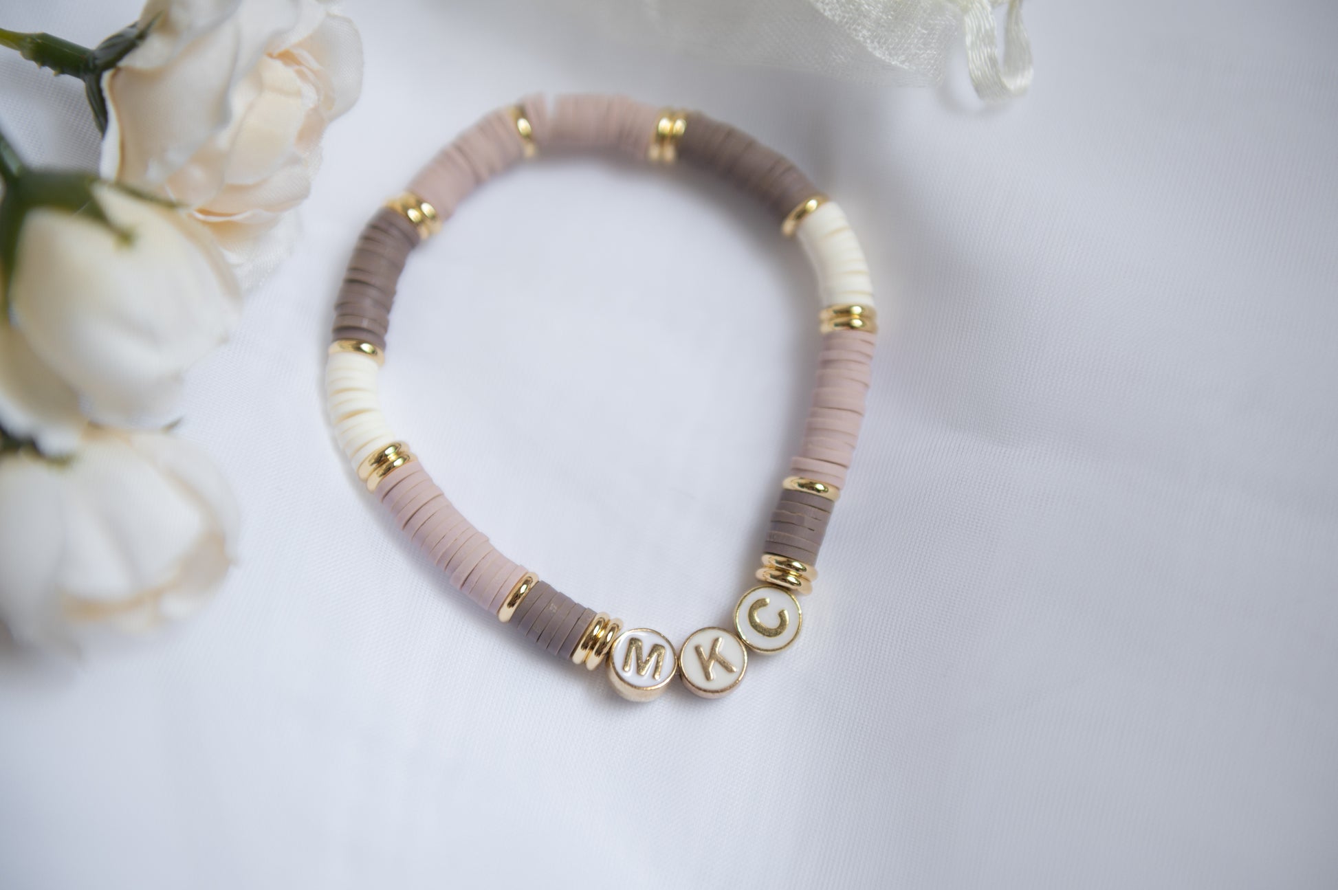 A clay-beaded bracelet with alternating dusty pink, taupe, and cream beads, accented with gold spacer rings and three round gold letter beads spelling baby's initials, displayed on a white surface with soft white flowers in the corner.