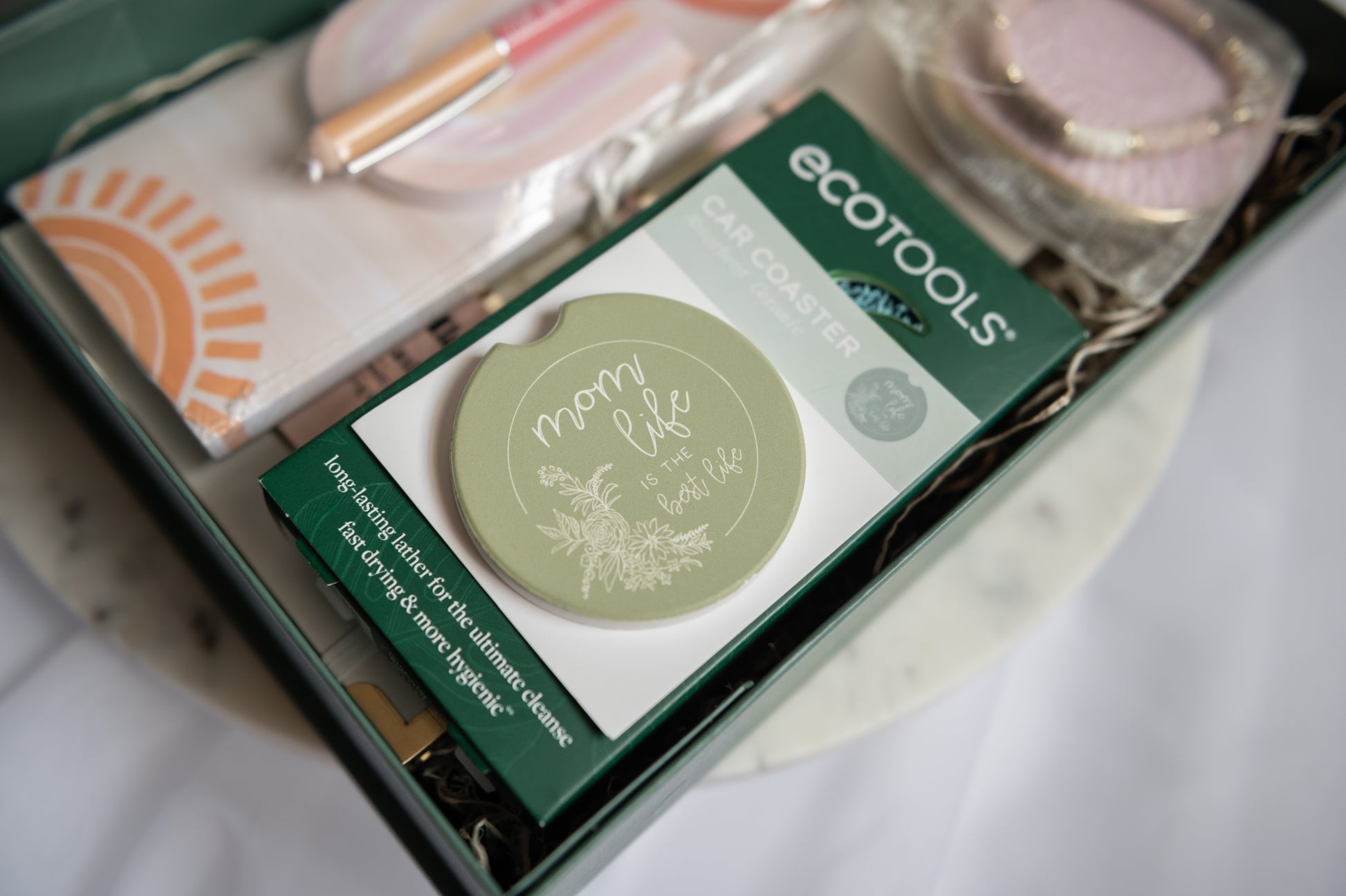 A gift box containing self care items for a postpartum mom heading back to work after maternity leave, including a round sage green coaster with 'mom life is the best life' printed on it, and other gift items in a styled arrangement.