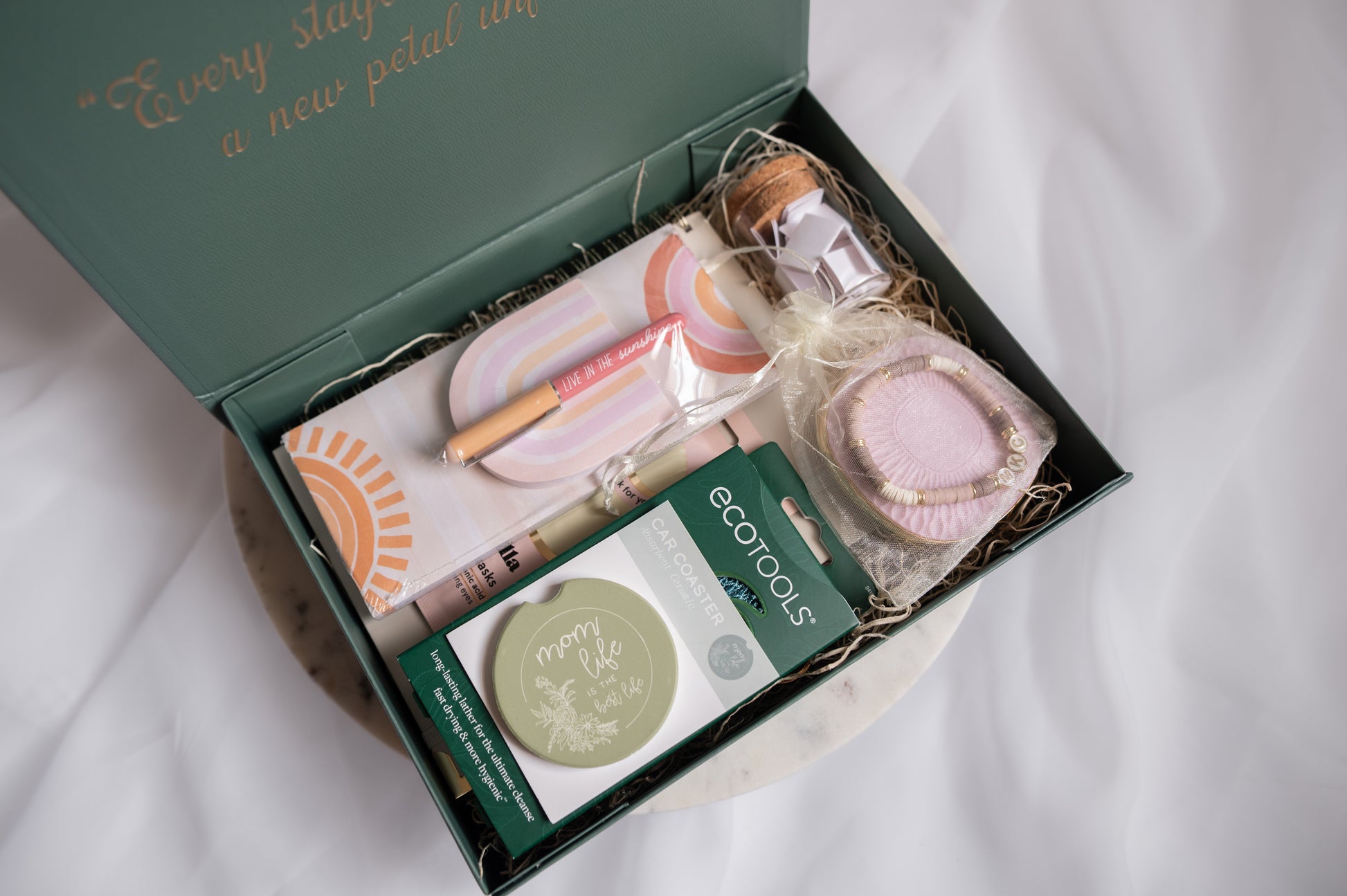 An open forest green gift box filled with spa and self-care items, including a postpartum journal, notepad set, a clay-beaded personalize bracelet with baby's initials, eye masks, and a round 'mom life' coaster, arranged on a white fabric background.