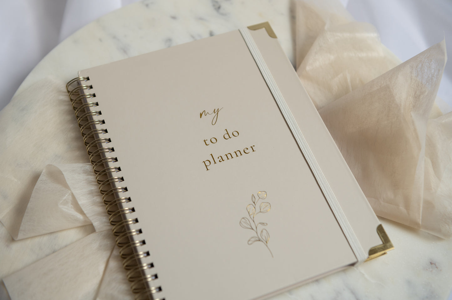 A cream-colored spiral-bound to-do planner with gold foil lettering and corner accents, styled on a marble surface with sheer cream fabric.