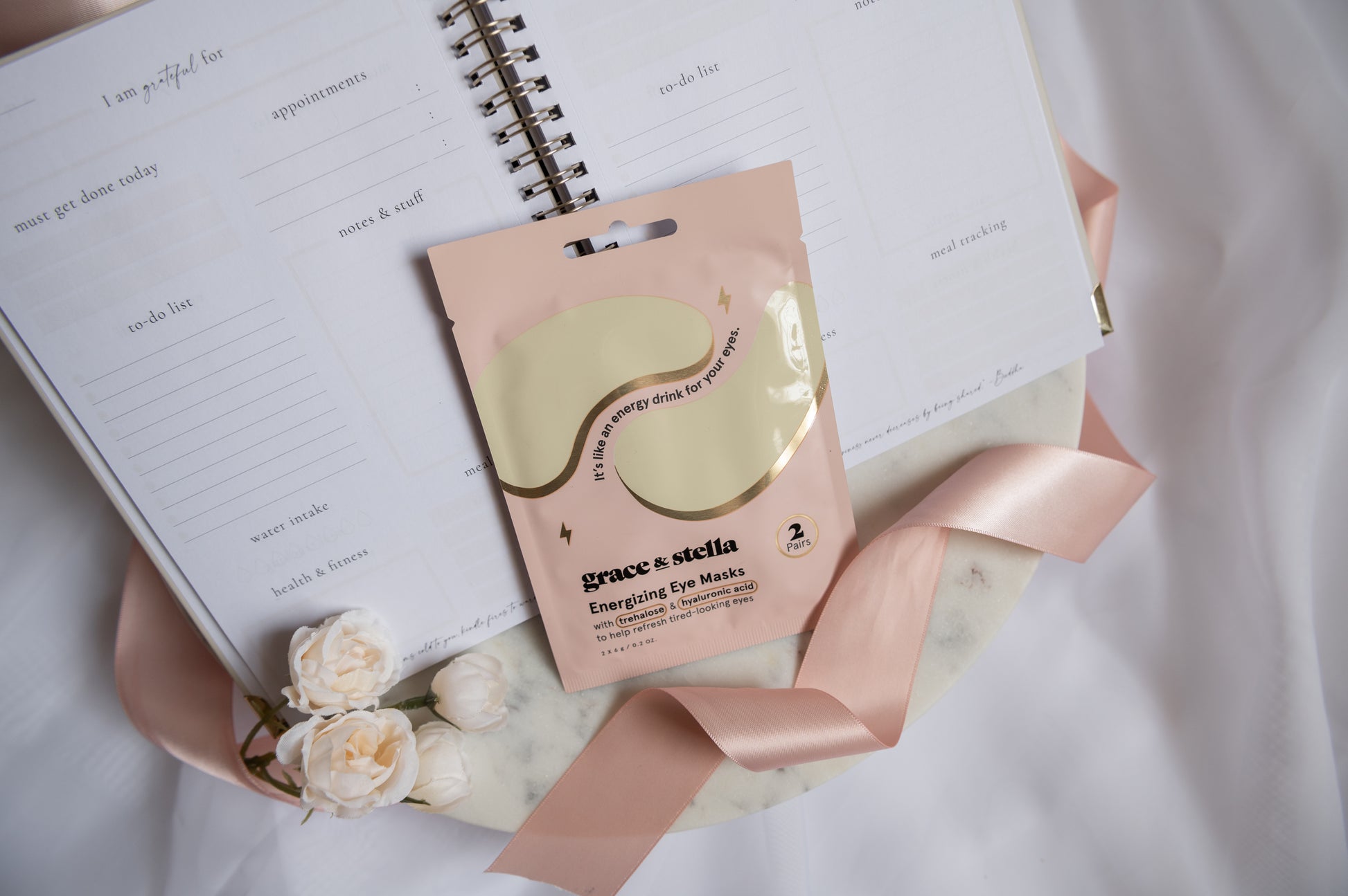 Grace & Stella Emerging Eye Masks in a pink pouch with gold foil detailing, styled on a marble coaster with a pink ribbon, cream roses, and an open planner.