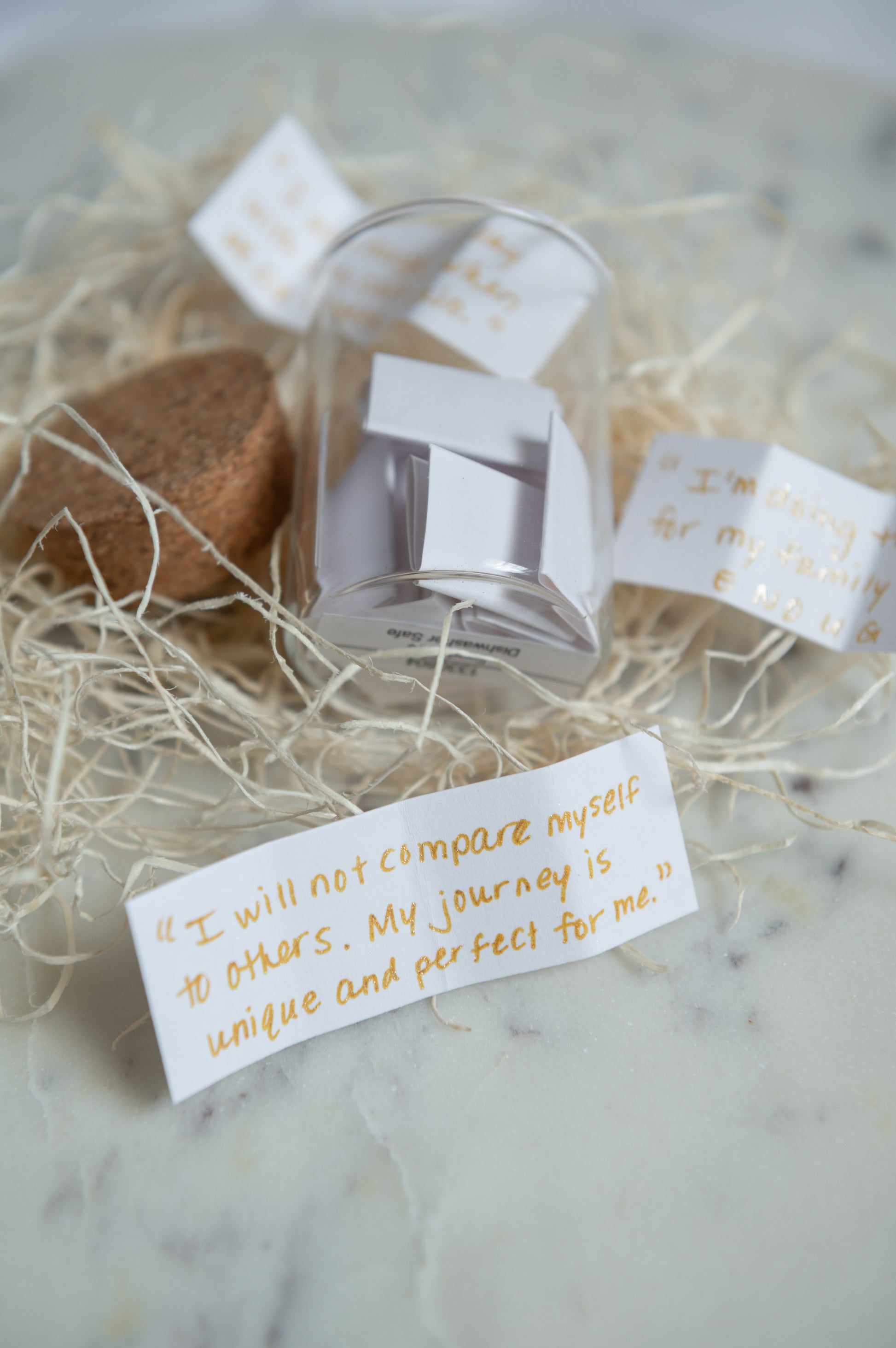 A clear cylindrical glass jar filled with folded white paper, placed on a marble-like surface with handwritten quotes and mom affirmations scattered nearby.