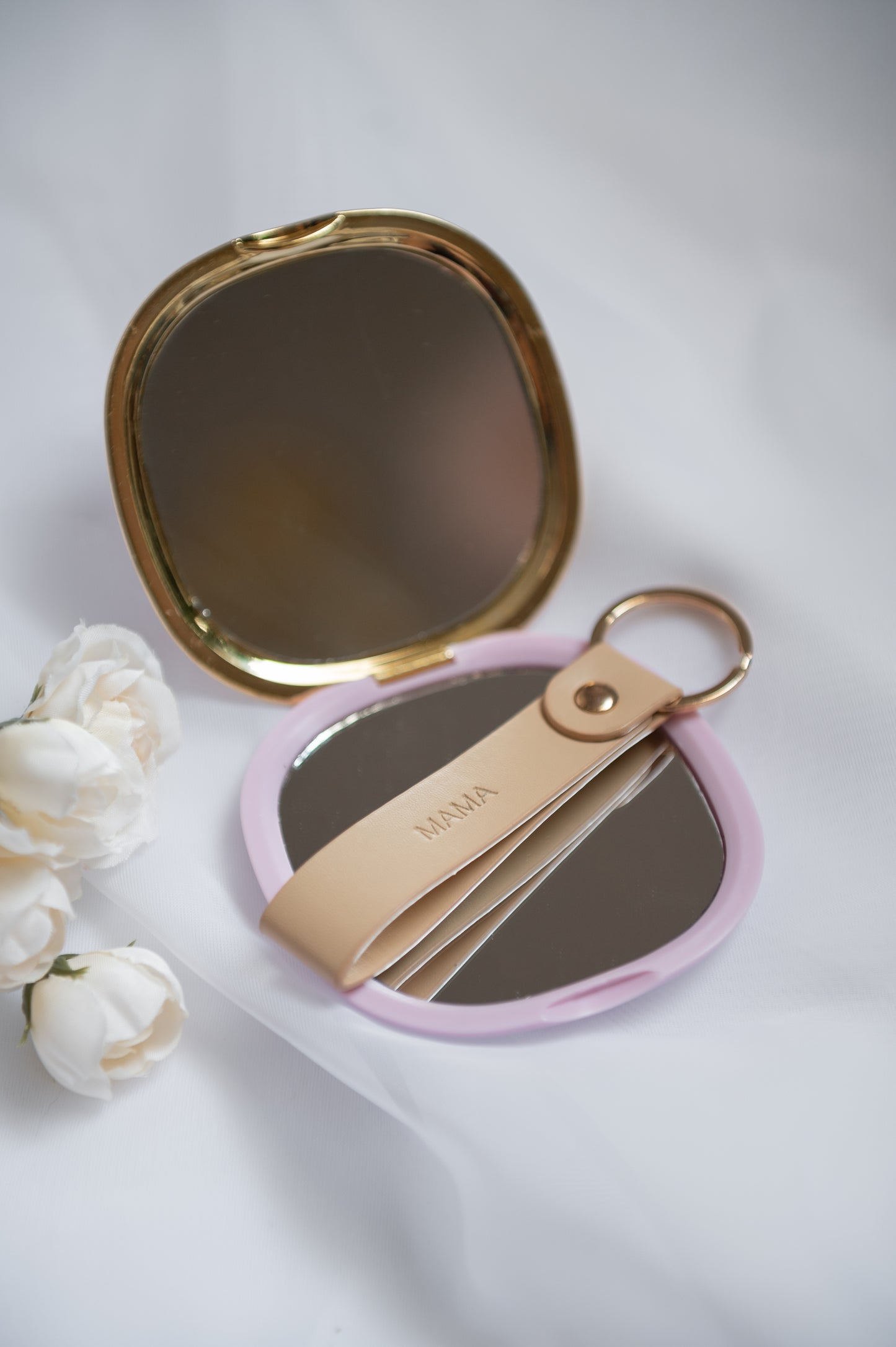 A compact folding mirror with a gold-tone frame, shown open with a light pink outer cover and a beige leather keychain with 'MAMA' embossed on the strap.