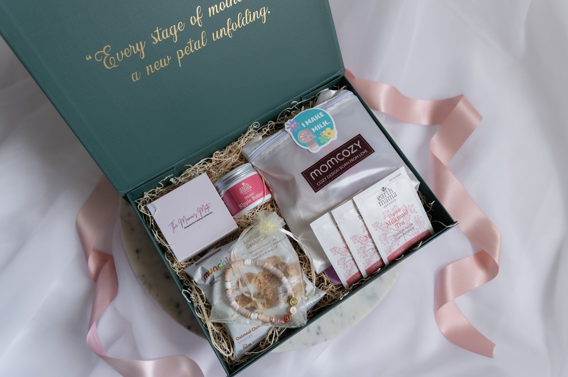 An open gift set filled with curated self-care and breastfeeding items, including nursing products, a personalized beaded bracelet, and 'The Mama's Mate' necklace, arranged in magnetic lid box on a white fabric background with a pink ribbon.
