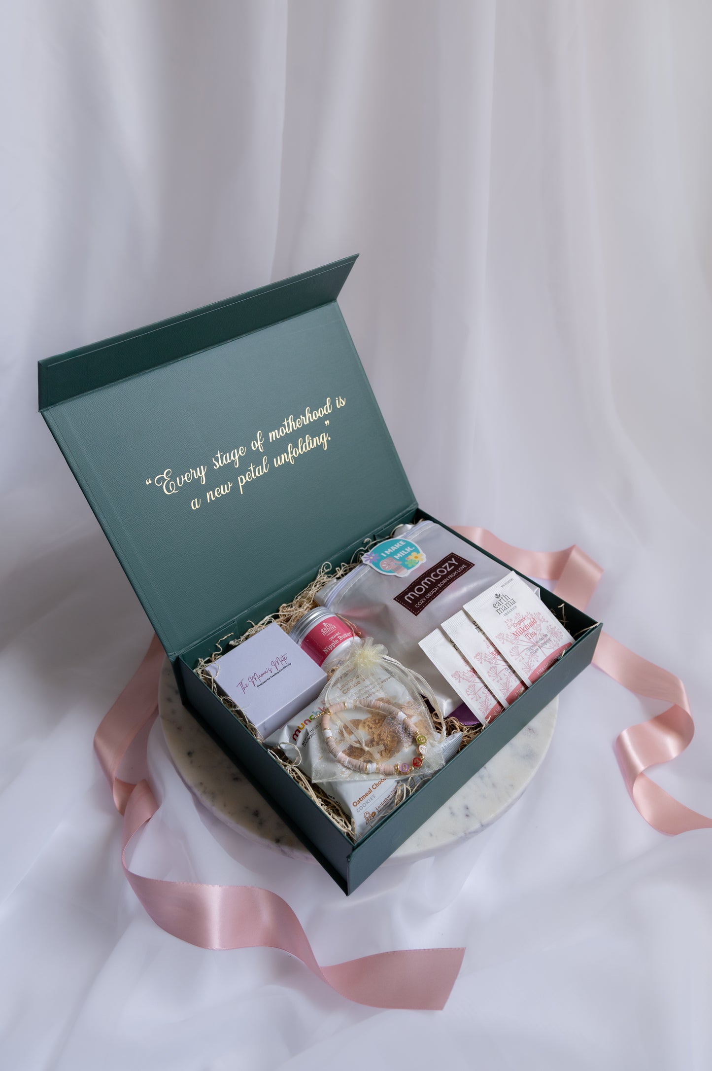 An forest green gift box with gold script on the inside lid, displayed on a white draped fabric background with a pink ribbon. The box contains self care items for a nursing or pumping mom, as well as other items to help her feel supported during her postpartum journey.
