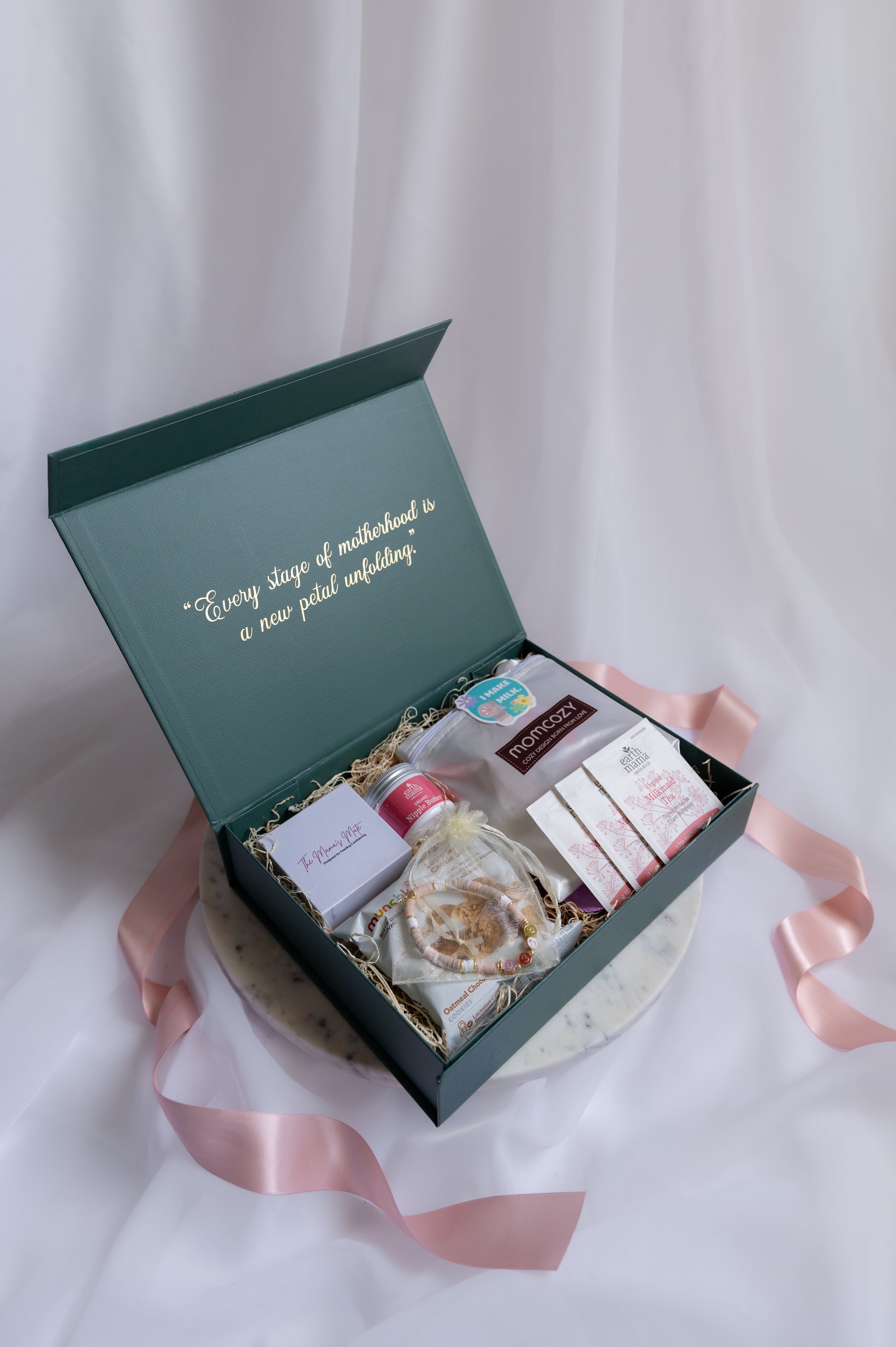 An forest green gift box with gold script on the inside lid, displayed on a white draped fabric background with a pink ribbon. The box contains self care items for a nursing or pumping mom, as well as other items to help her feel supported during her postpartum journey.