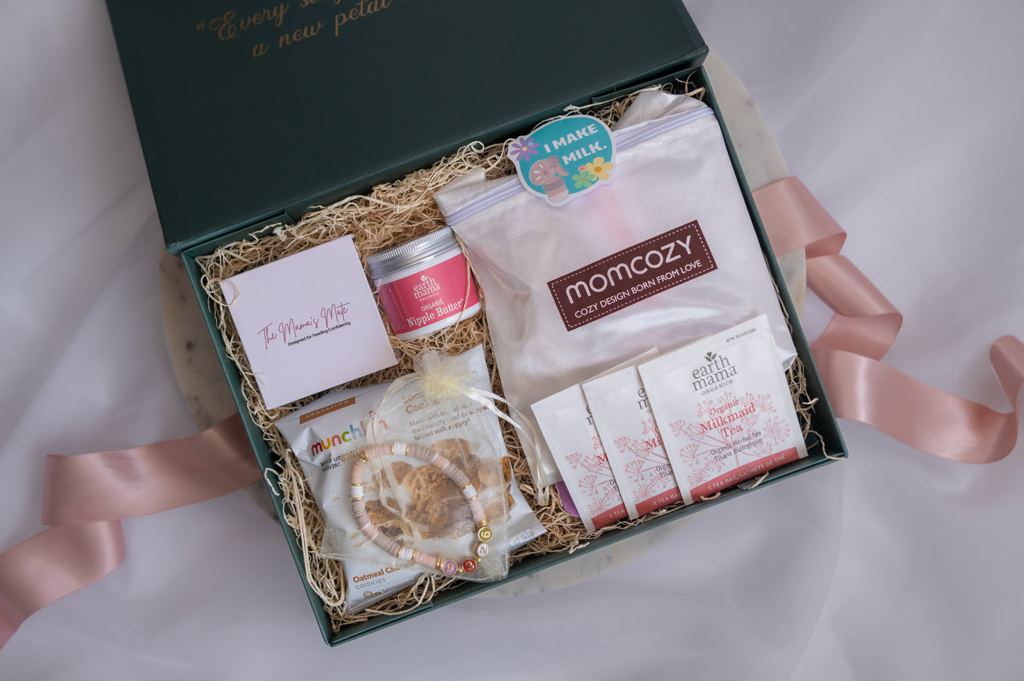 An open dark green gift box filled with baby care and mom-and-me treats on a white fabric background. The box contains a pink card, a small pink tin, a clear bag of Momcozy baby milk powder, packaged snacks, and a beaded bracelet, tied with a pale pink ribbon.