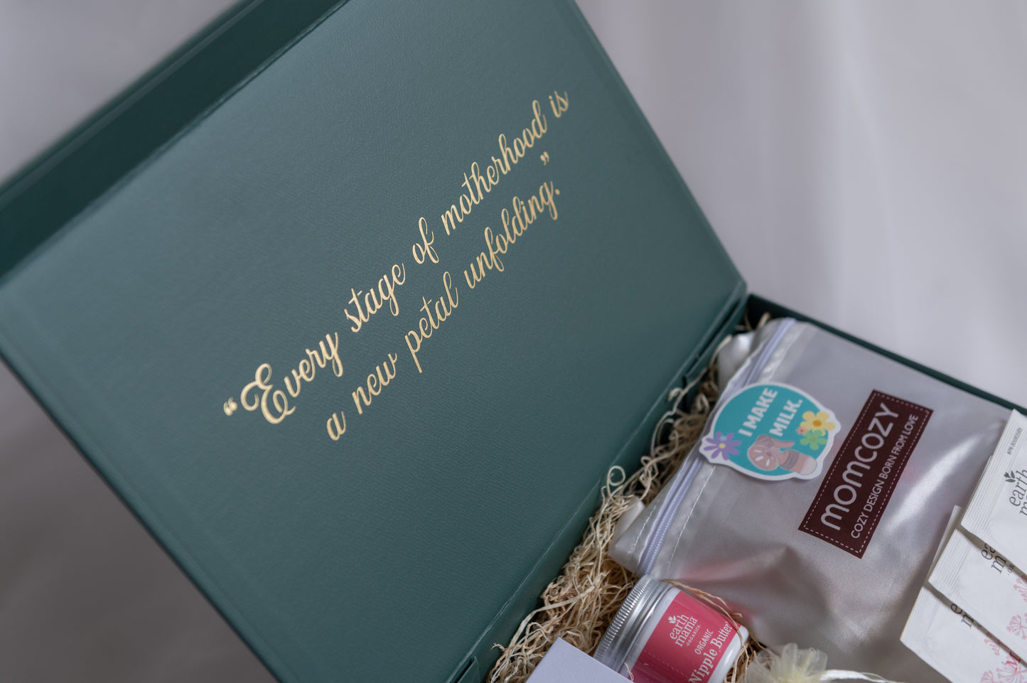 An open forest grean gift box with gold foil text reading, "Every stage of motherhood is a new petal unfolding." Inside, a Momcozy nursing pads, Earth Mama nipple butter, and other breastfeeding/pumping items are arranged on luxury filler against a soft light background.