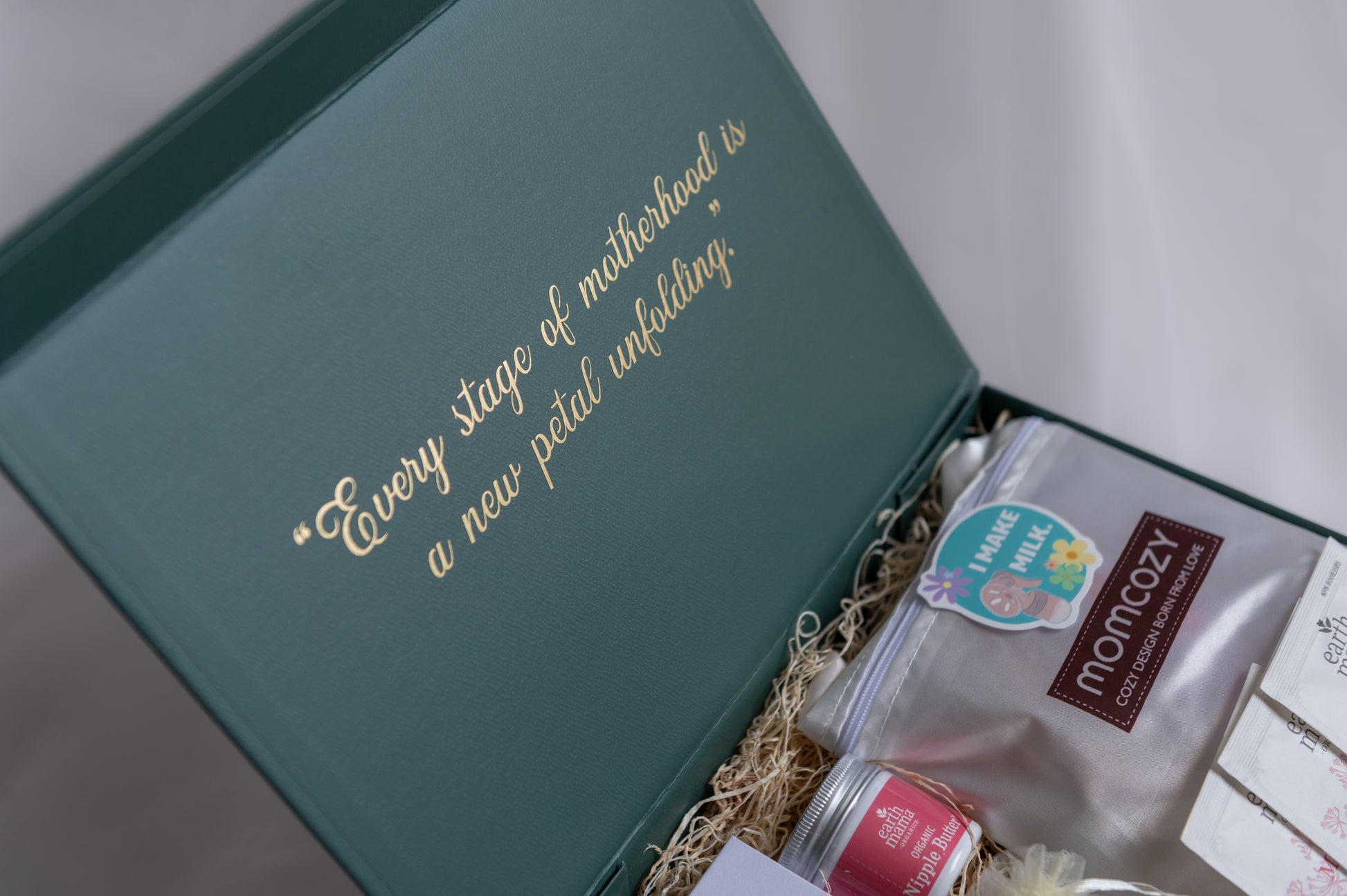 An open forest grean gift box with gold foil text reading, "Every stage of motherhood is a new petal unfolding." Inside, a Momcozy nursing pads, Earth Mama nipple butter, and other breastfeeding/pumping items are arranged on luxury filler against a soft light background.