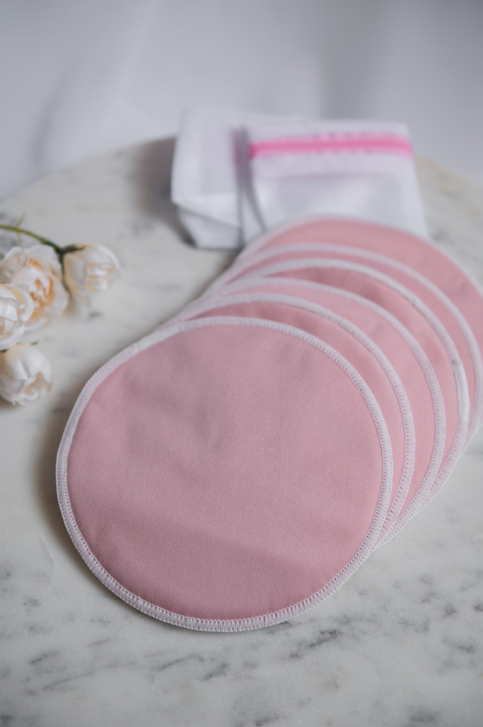 Six pink reusable nursing or breastfeeding pads with white trim arranged in a fan shape on a white marble surface, with a folded white cloth and pink zipper pouch in the background.