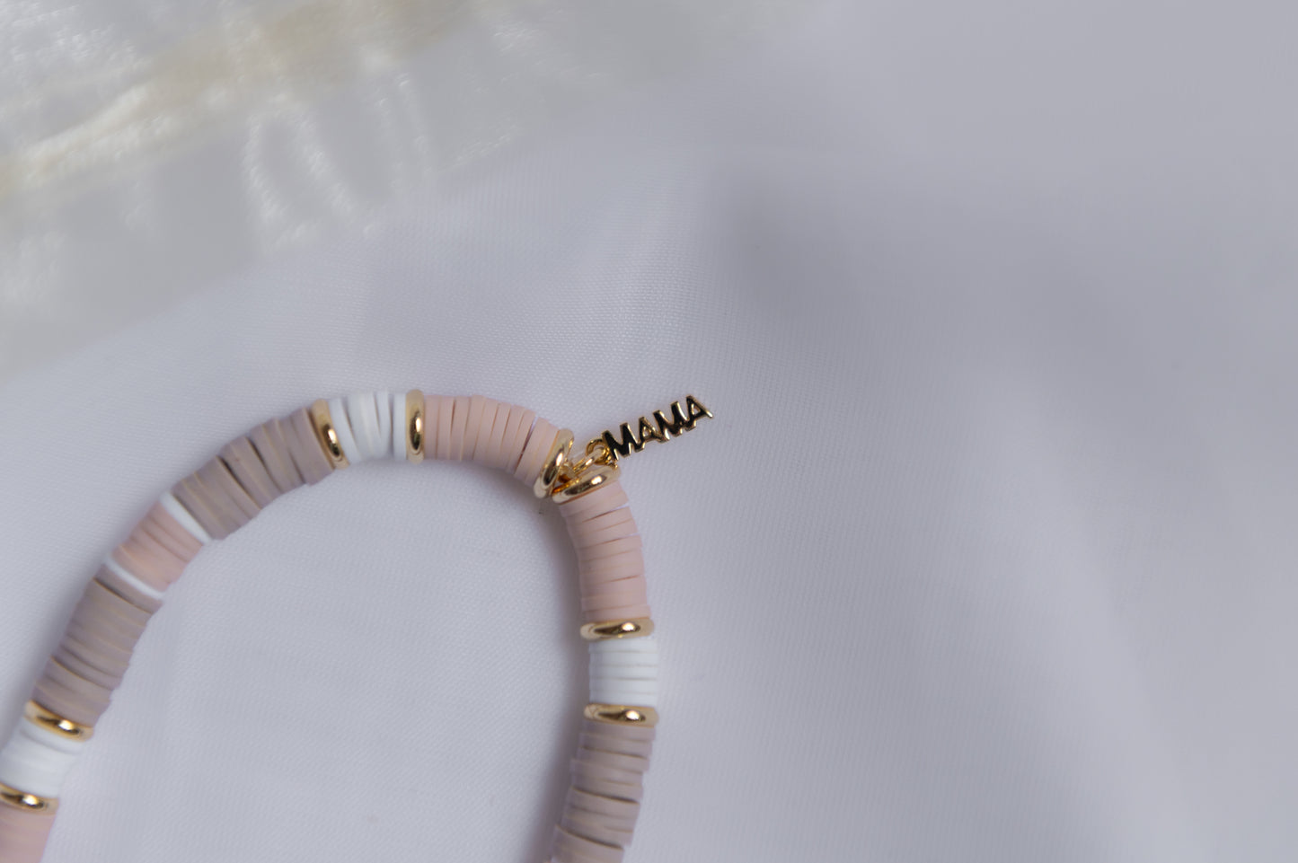 A close-up of a clay-beaded bracelet with alternating light pink, white, and taupe-gray discs, accented with small gold spacer beads and a gold-tone 'MAMA' script charm.