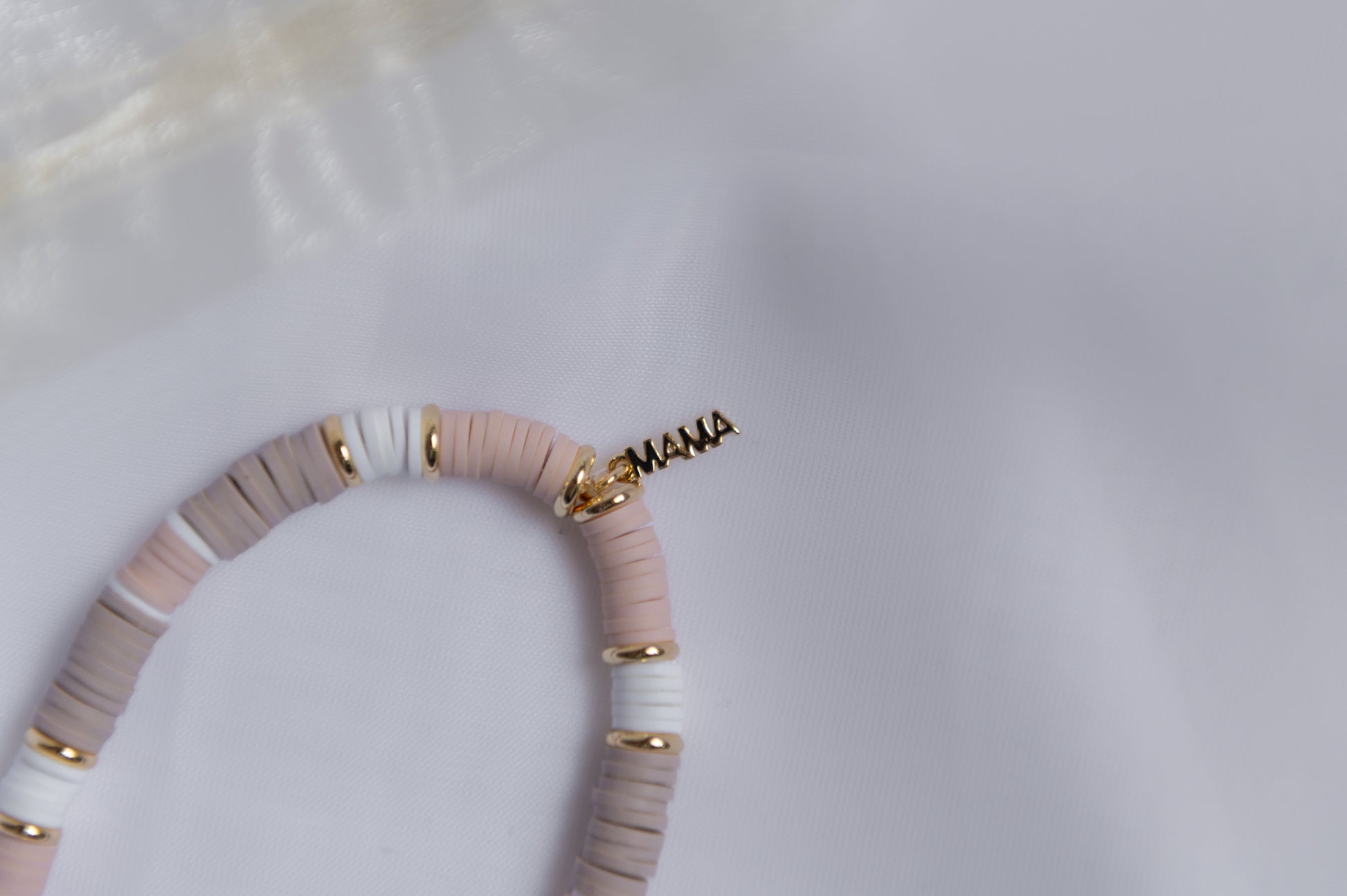A close-up of a clay-beaded bracelet with alternating light pink, white, and taupe-gray discs, accented with small gold spacer beads and a gold-tone 'MAMA' script charm.