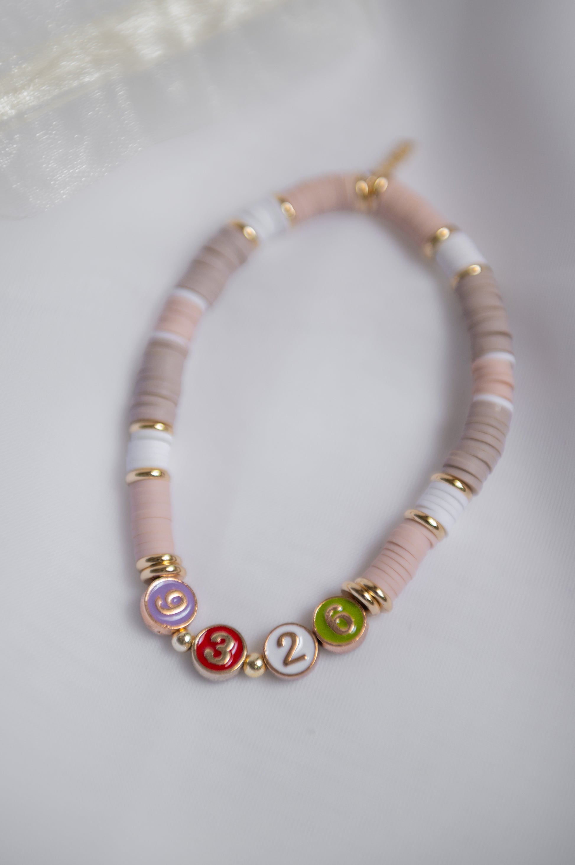 A clay beaded necklace, alternating light pink and taupe-gray beads, accented with white beads and gold spacer rings. The front features three round enamel charms in purple, red, and green with gold lettering, displayed on a soft white fabric background.