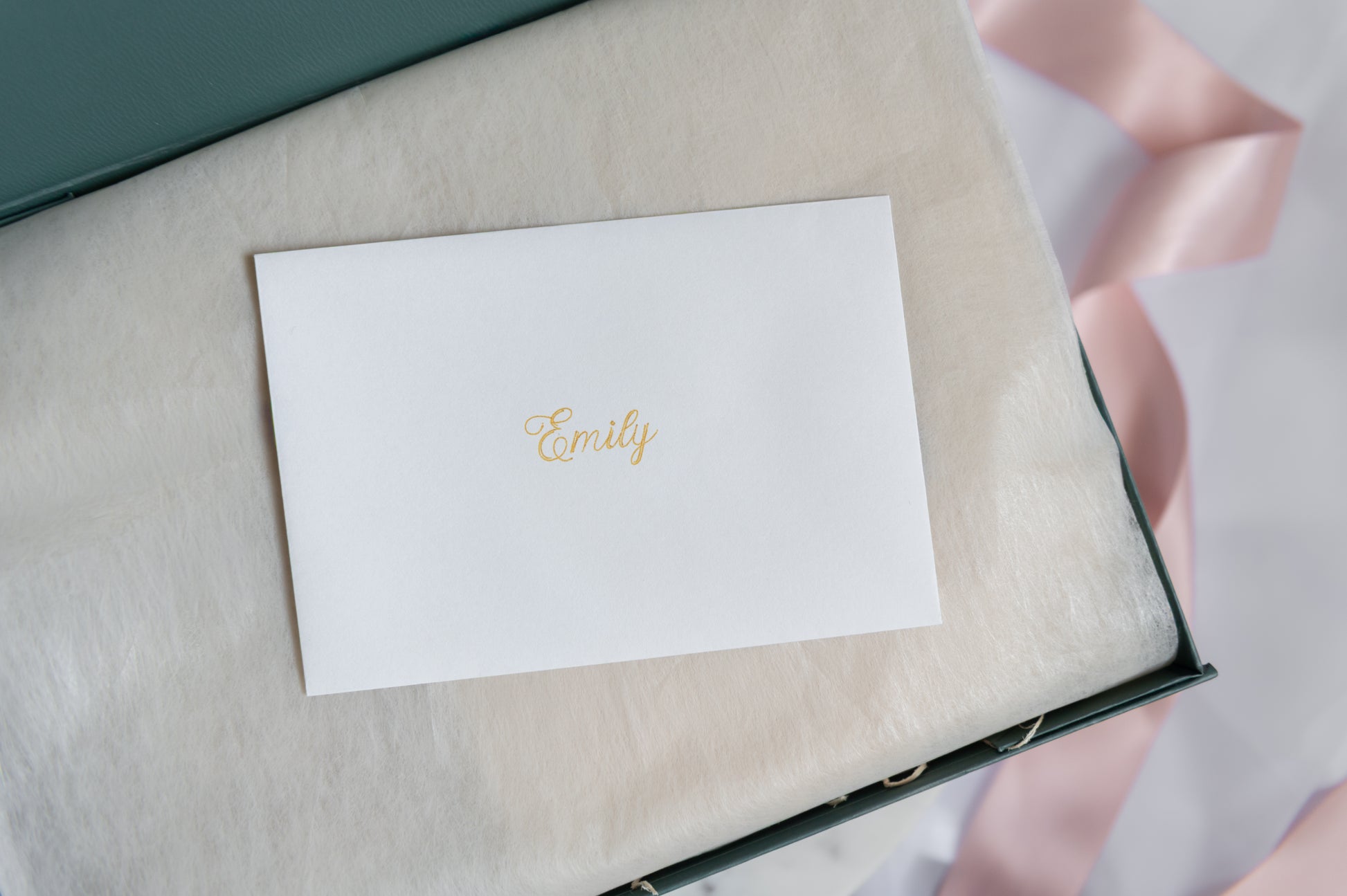 A white personalized card with the recipient's name in gold foil script, placed on a soft cream-colored insert inside a dark green gift box, with a pale pink ribbon in the background.