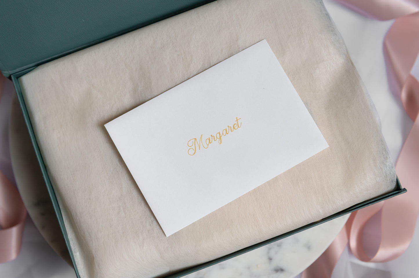 A white card with the recipient's name in gold cursive script, placed on a beige tissue insert inside a dark green gift box, with pink satin ribbons around it.