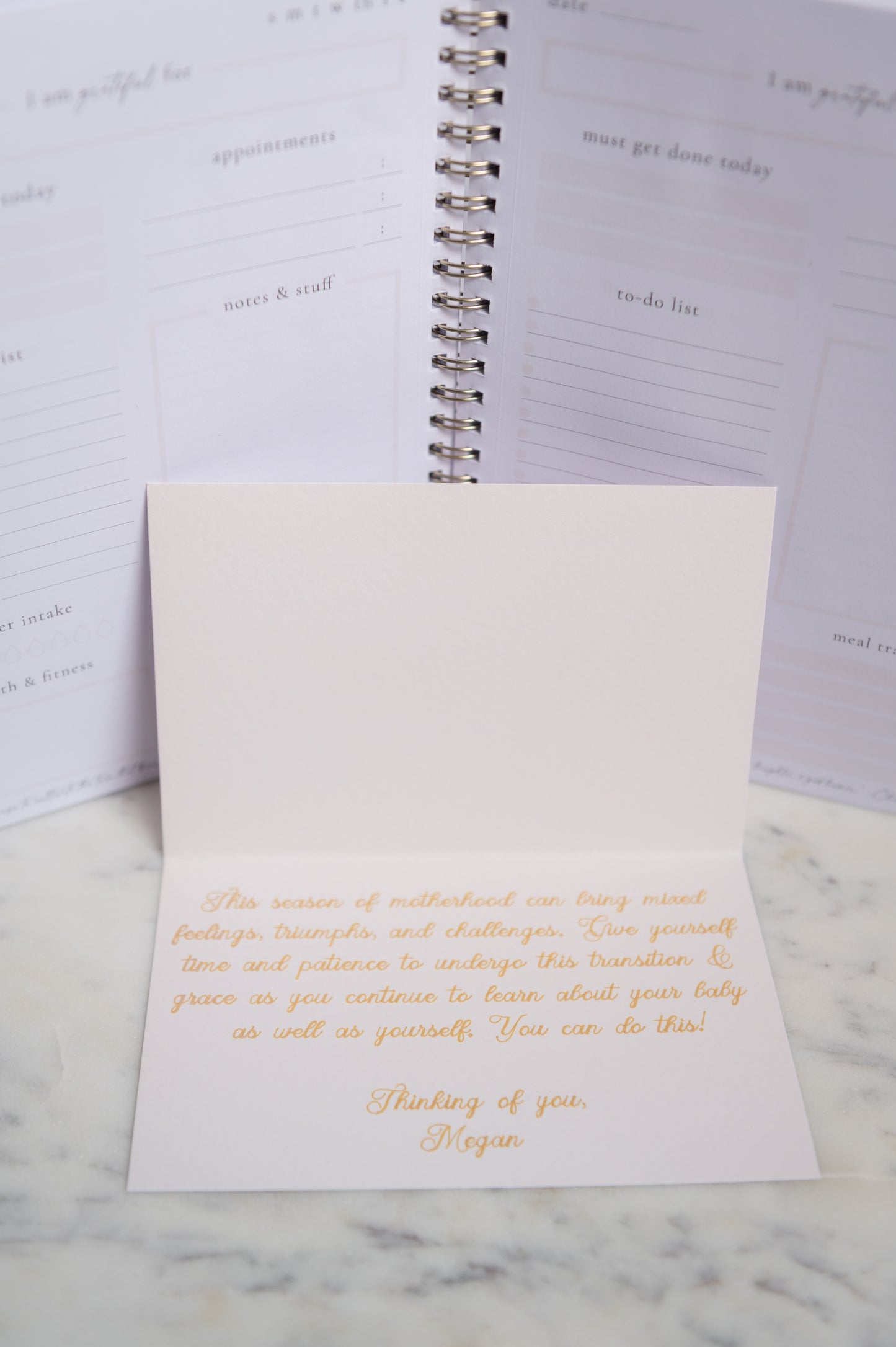 A square cream greeting card with elegant gold foil lettering stands in front of an open spiral notebook on a light marble surface.