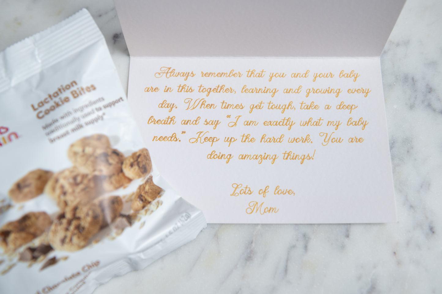 An open white greeting card with gold cursive text lies on a light marble surface, next to a bag of lactation cookie bites. The card is the main product being shown, with the snack bag acting as a prop.