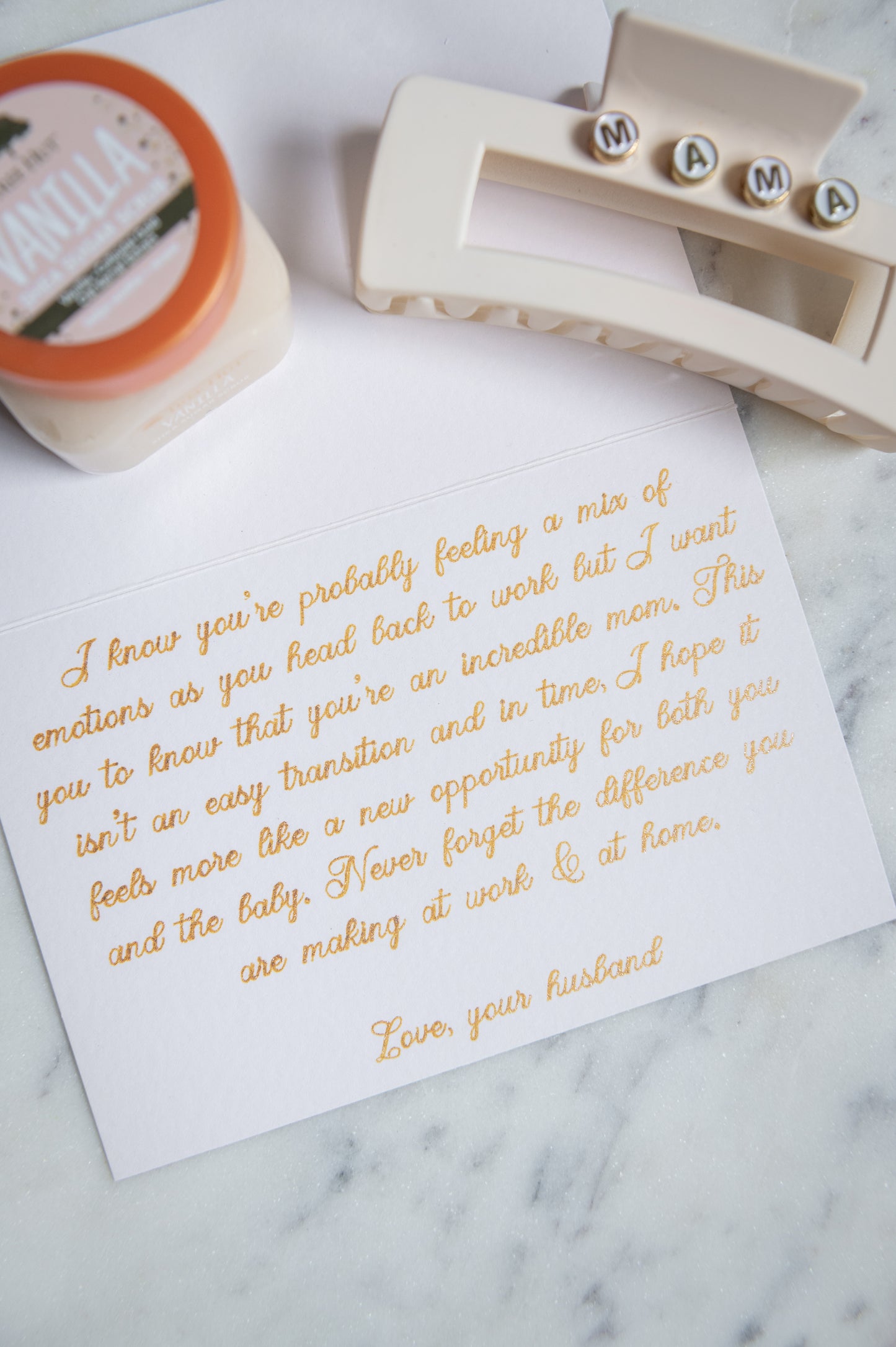 A white square card with elegant gold foil script text, styled on a light marble surface alongside a cream hair claw clip and a small jar of vanilla body scrub.