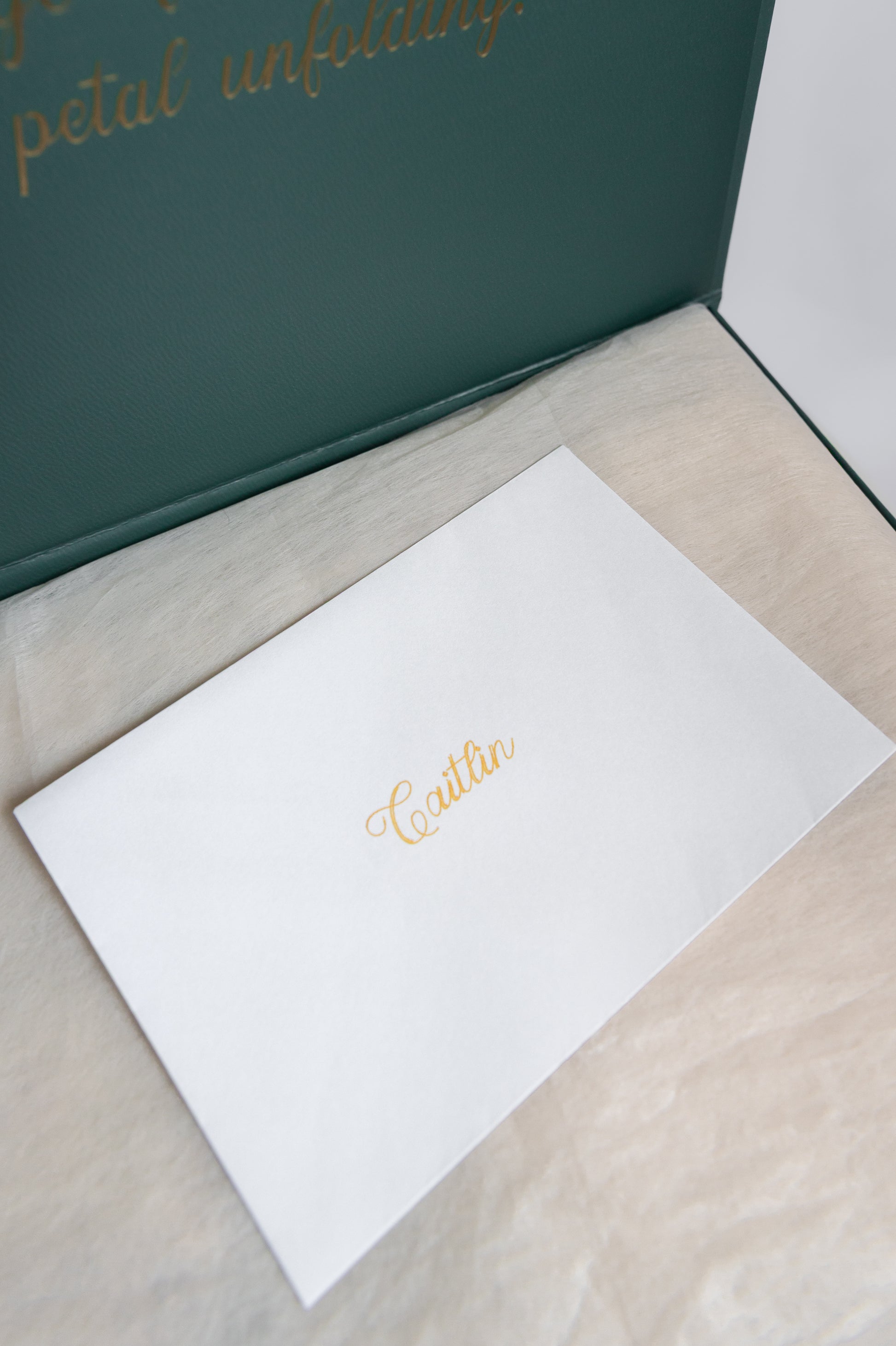 A white envelope with the recipient's name in gold cursive script, placed on a light beige textured surface next to a dark green box lid with gold lettering.